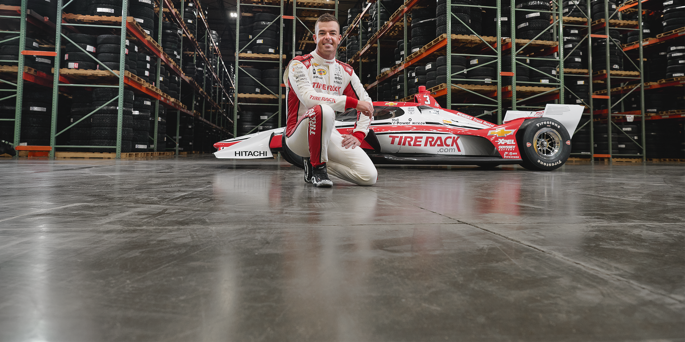 cott McLaughlin kneeling in front of his Team Penske IndyCar at the Tire Rack warehouse, promoting the Tire Rack sponsorship ahead of the 2025 Detroit Grand Prix
