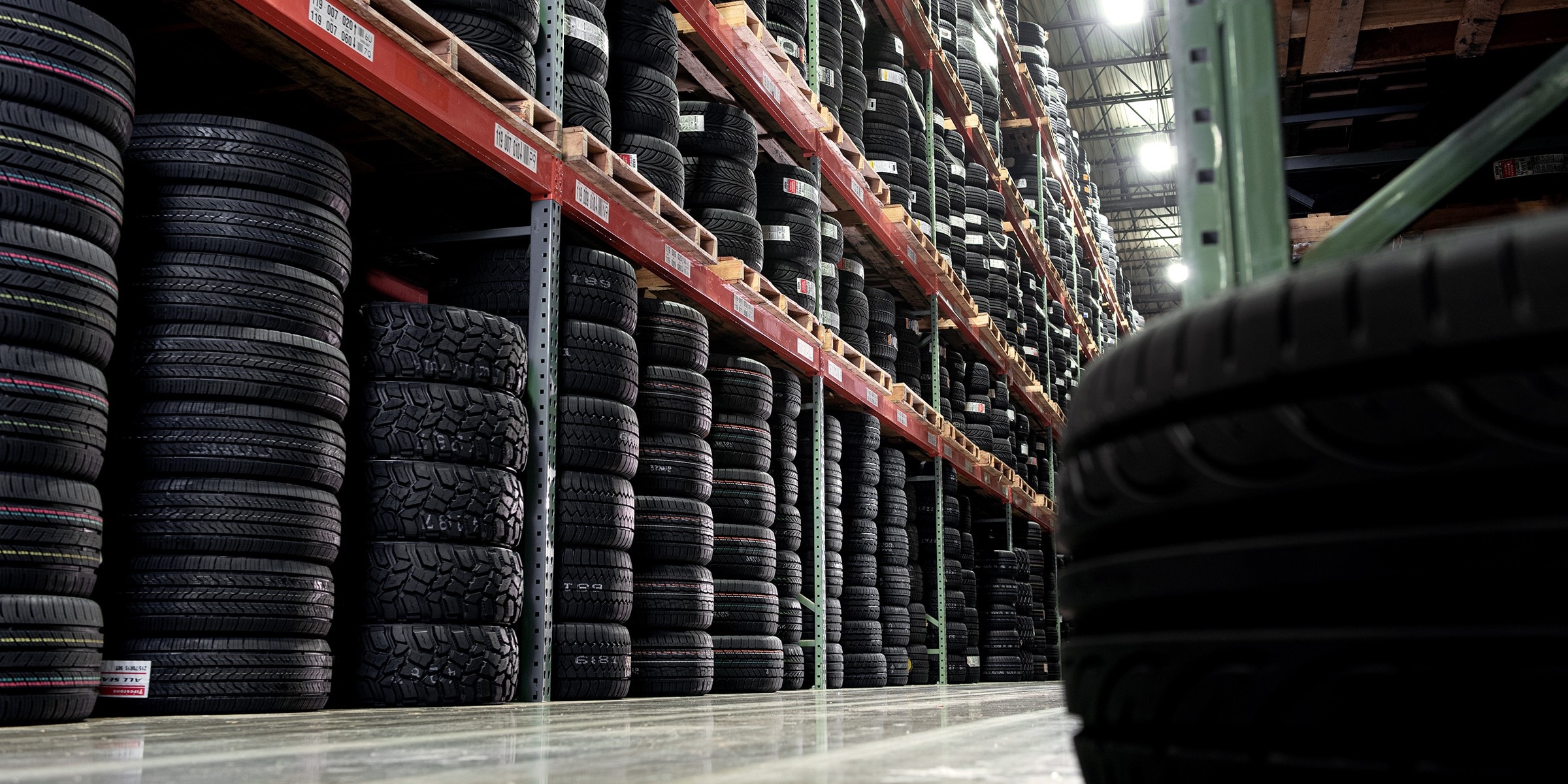 Stacks of tires and wheels in a warehouse aisle, representing the wide selection, expert reviews, and support Tire Rack offers to help you get there.