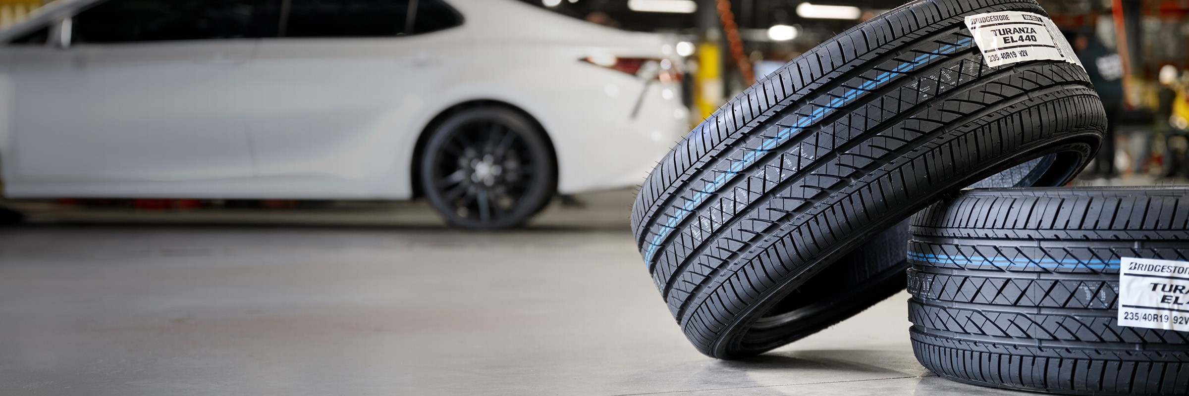 New pair of Bridgestone Turanza tires in a service bay with a white sedan in the background.
