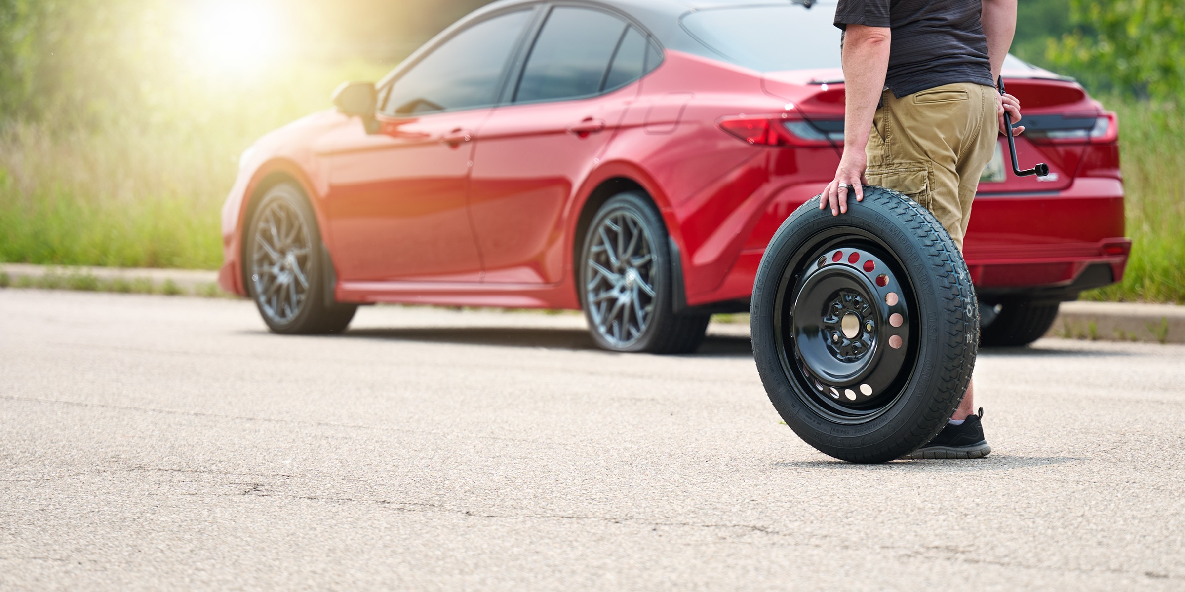 Driver rolling a special use spare tire toward a red sedan on the roadside.